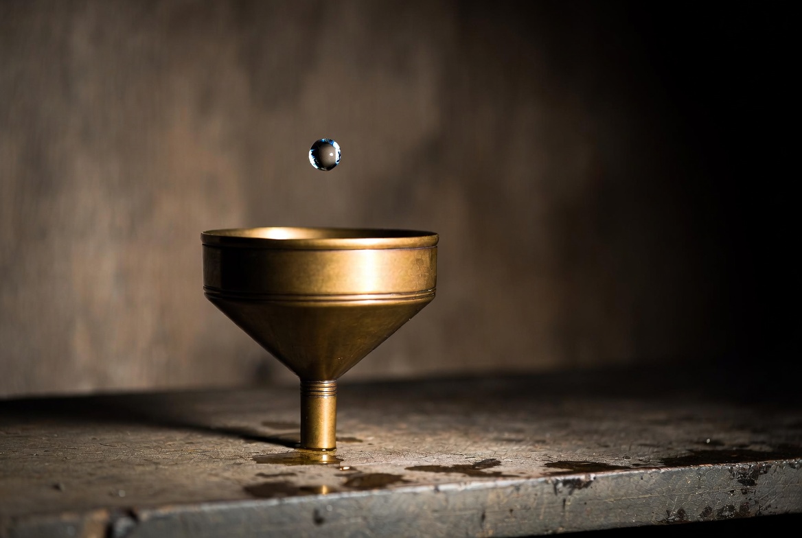 A single brass funnel on a worn wooden workbench with one droplet caught mid-fall above its narrow neck, sharp focus on the droplet, soft side light, no people, editorial.