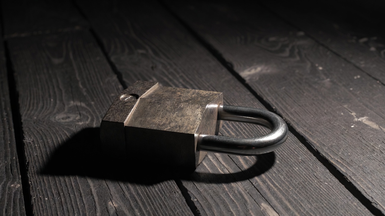 A heavy steel padlock lying open on a dark wooden surface with no key visible, hard overhead light casting a sharp shadow.