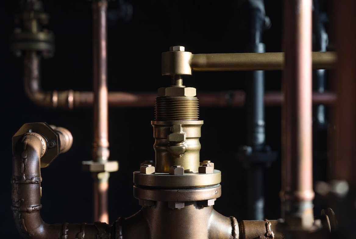 A single brass valve mid-flow on a polished copper pipe assembly against a charcoal background, sharp side lighting catching the valve's machined edges, no people, editorial.