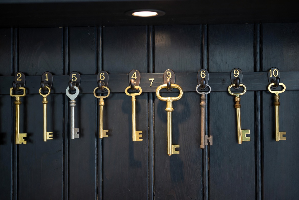 A row of brass keys hanging on numbered hooks against a dark wooden panel wall, one hook empty with a brighter outline, single overhead light, no people, editorial.