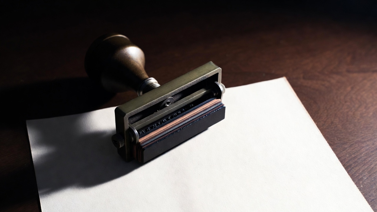 A heavy metal date stamp poised over a clean white document, ink visible on the stamp rubber, single overhead light.