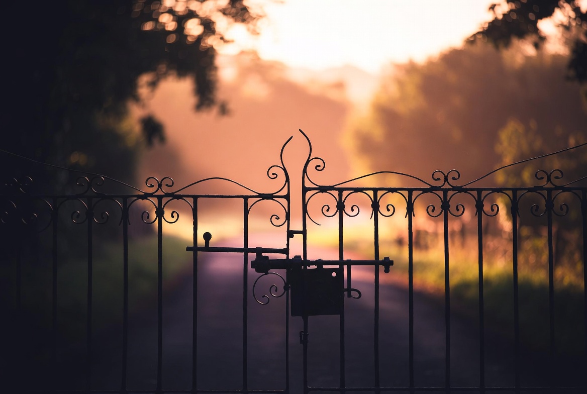 A wrought-iron gate half-open across a country lane at golden hour, sharp focus on the latch mechanism, soft warm light, no people, editorial.