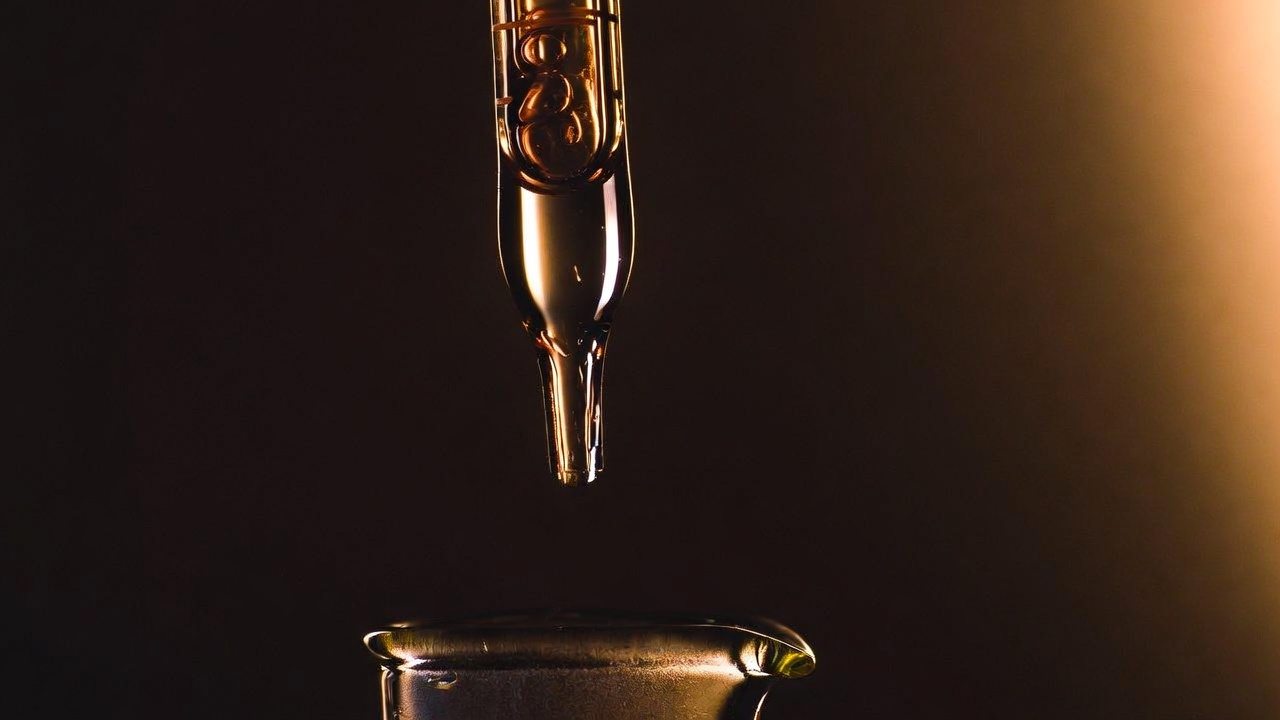 A clear glass laboratory pipette suspended over a beaker with a single amber drop forming at its tip.