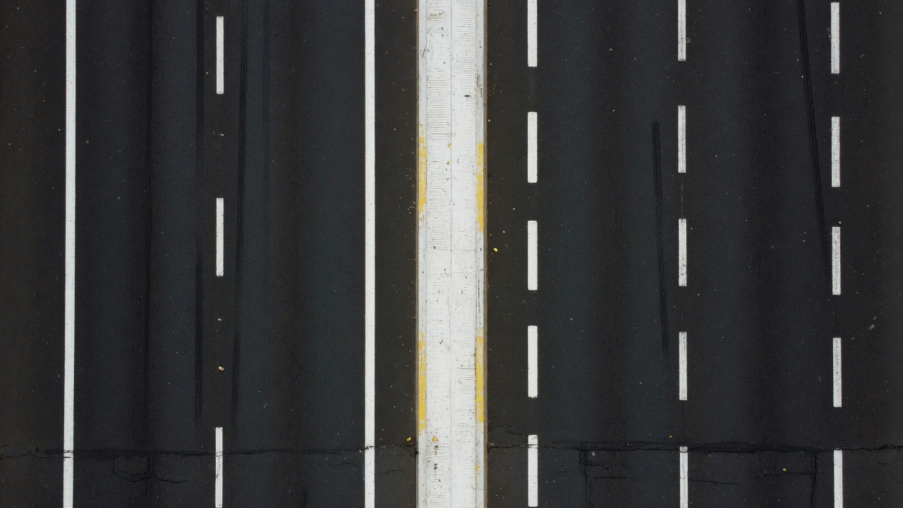 Two parallel road lanes on dark asphalt photographed straight from above — one marked with short dashed white paint, one marked with a continuous solid white line, a thick divider between them.