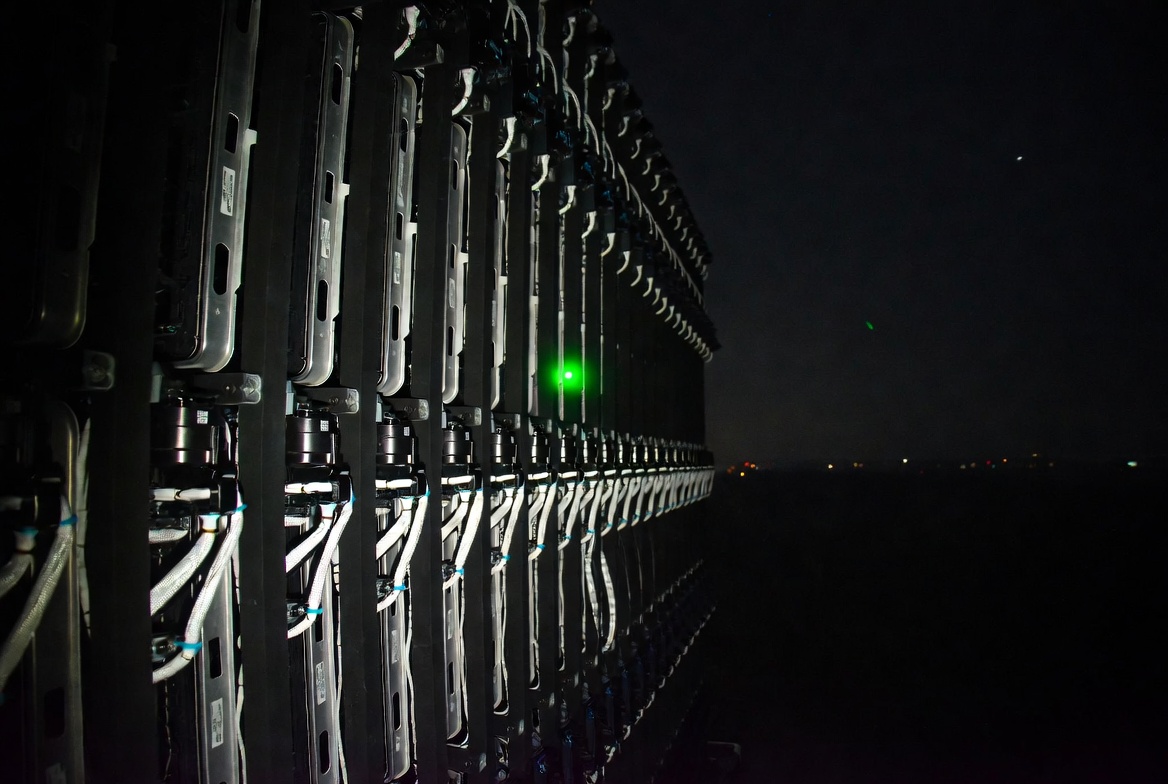 A black-and-white wide shot of a server rack at night with a single green LED illuminating one rack unit, cables draping like ivy down the side, no people, dramatic editorial composition.